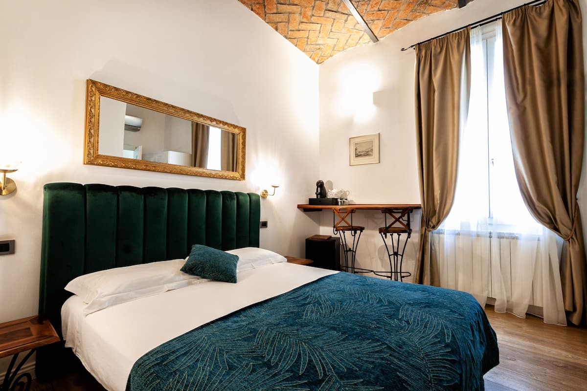 A hotel room with a green upholstered headboard, a large mirror, a bed with white linens and a blue patterned bedspread, a desk with a chair, and a window with beige curtains.