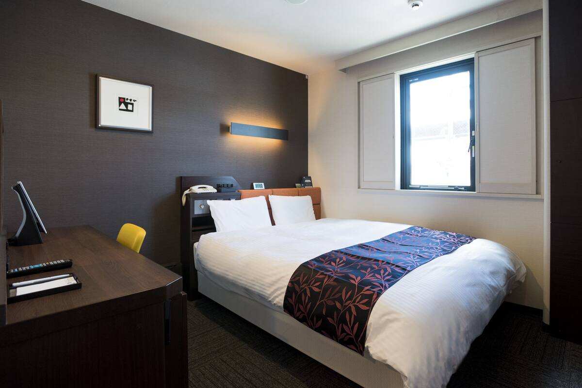 Une chambre d’hôtel comprenant un lit, un bureau avec un téléphone, une chaise, une fenêtre et un luminaire fixé au mur.