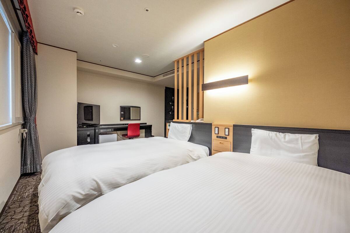 Une chambre d’hôtel avec deux lits, un bureau avec un ordinateur, une télévision et une lampe fixée au mur.