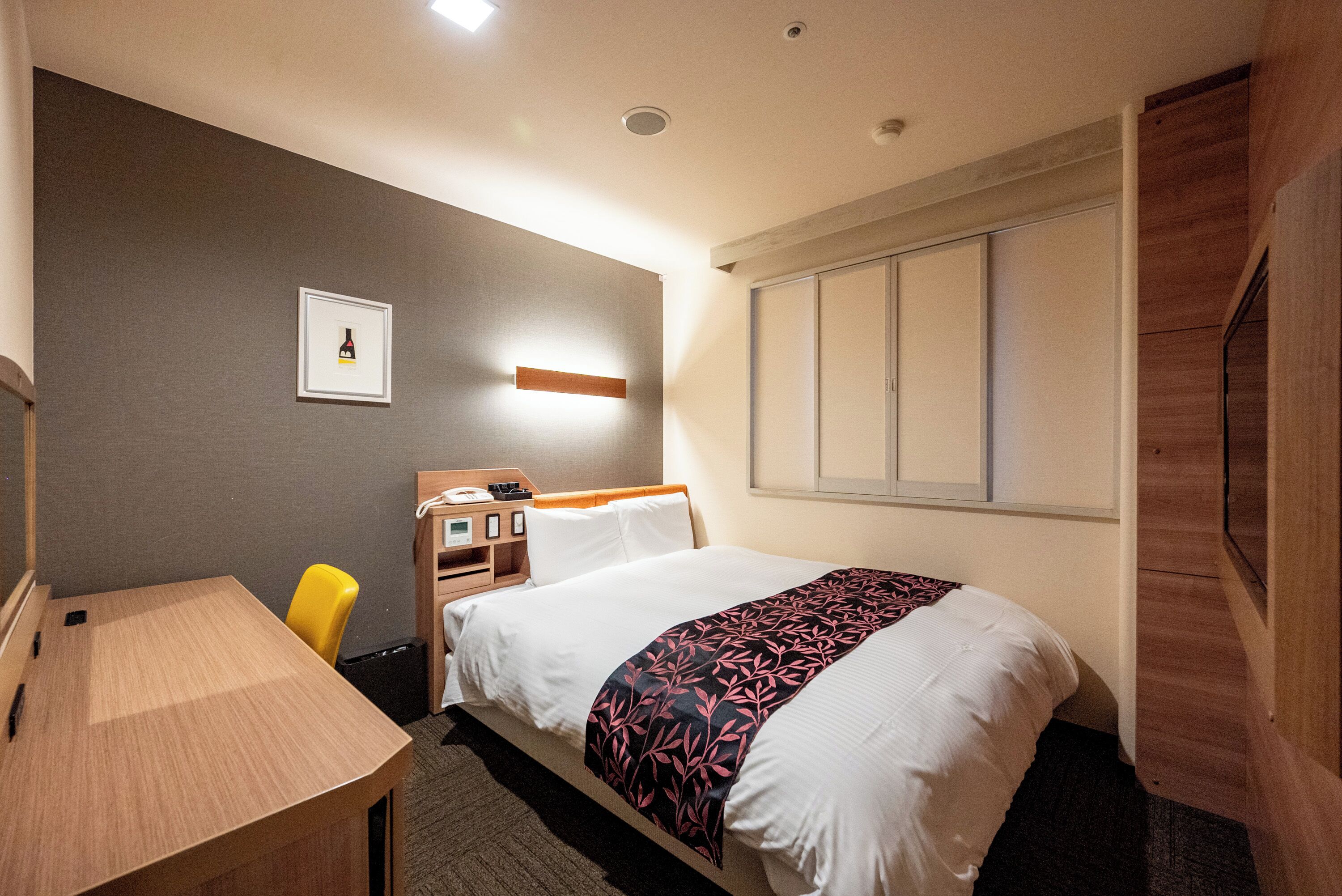 A hotel room with a bed, a desk, a chair, a bedside table, a nightstand, a framed picture on the wall, and a window with blinds.