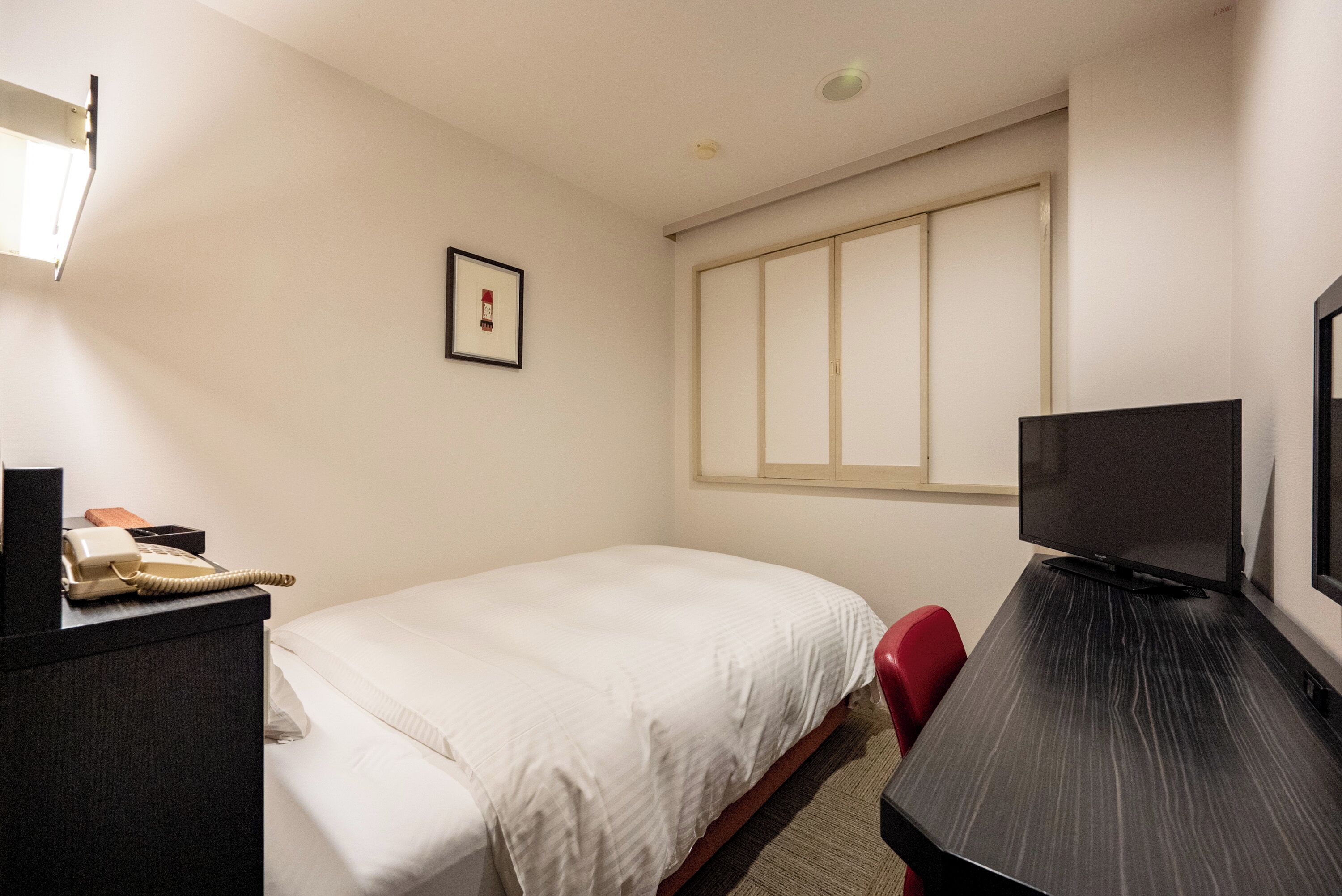 A hotel room with a bed, a desk with a television, a red chair, a telephone, and a framed picture on the wall.