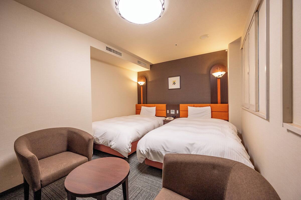 Une chambre d’hôtel avec deux lits simples, une petite table ronde, deux fauteuils et une lampe fixée au mur.