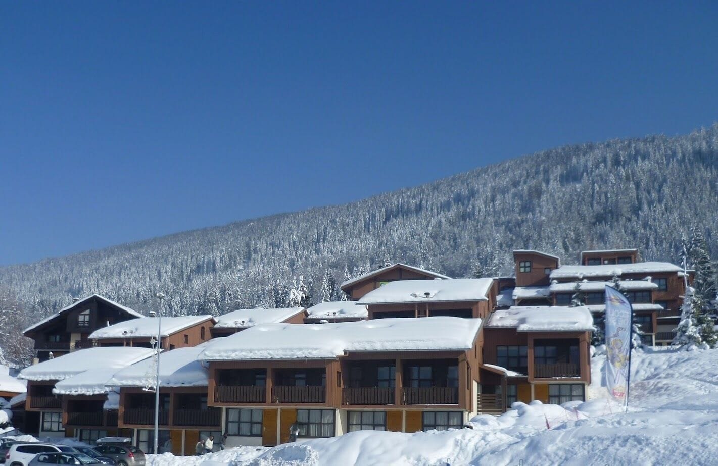A cluster of wooden cabins with snow-covered roofs, set against a backdrop of a snow-laden mountain.