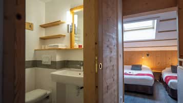 Una habitación con techo de madera, tragaluz, cama con manta roja, mesita de noche y baño con lavamanos e inodoro.