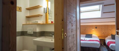 Ein Zimmer mit Holzbalkendecke, einem Oberlicht, einem Bett mit roter Decke, einem Nachttisch und einem Badezimmer mit Waschbecken und Toilette.