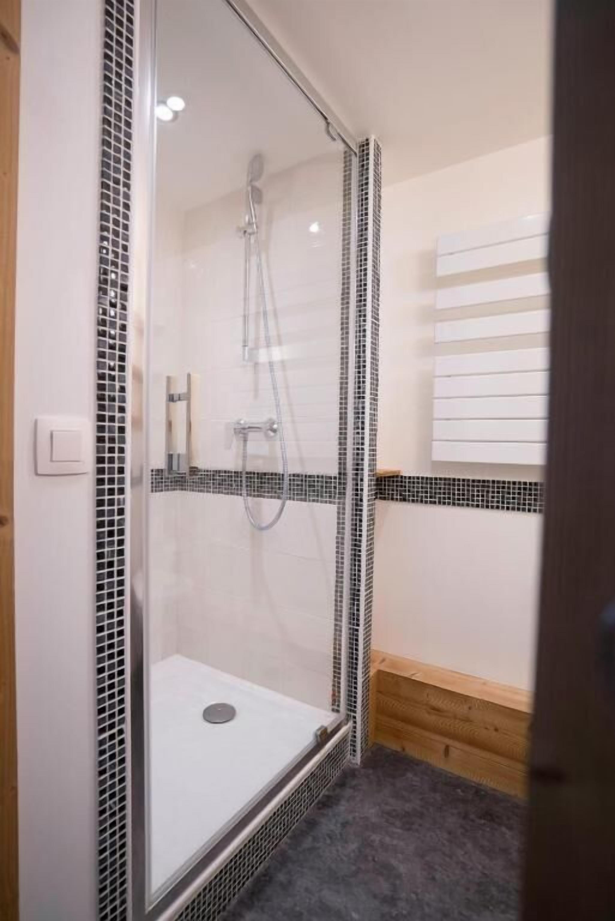 Ducha con cerramiento de vidrio, borde de azulejos, banco de madera y toallero blanco.