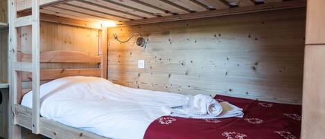 Una cama litera de madera individual con una colcha roja y ropa de cama blanca, una lámpara fijada a la pared y un enchufe.
