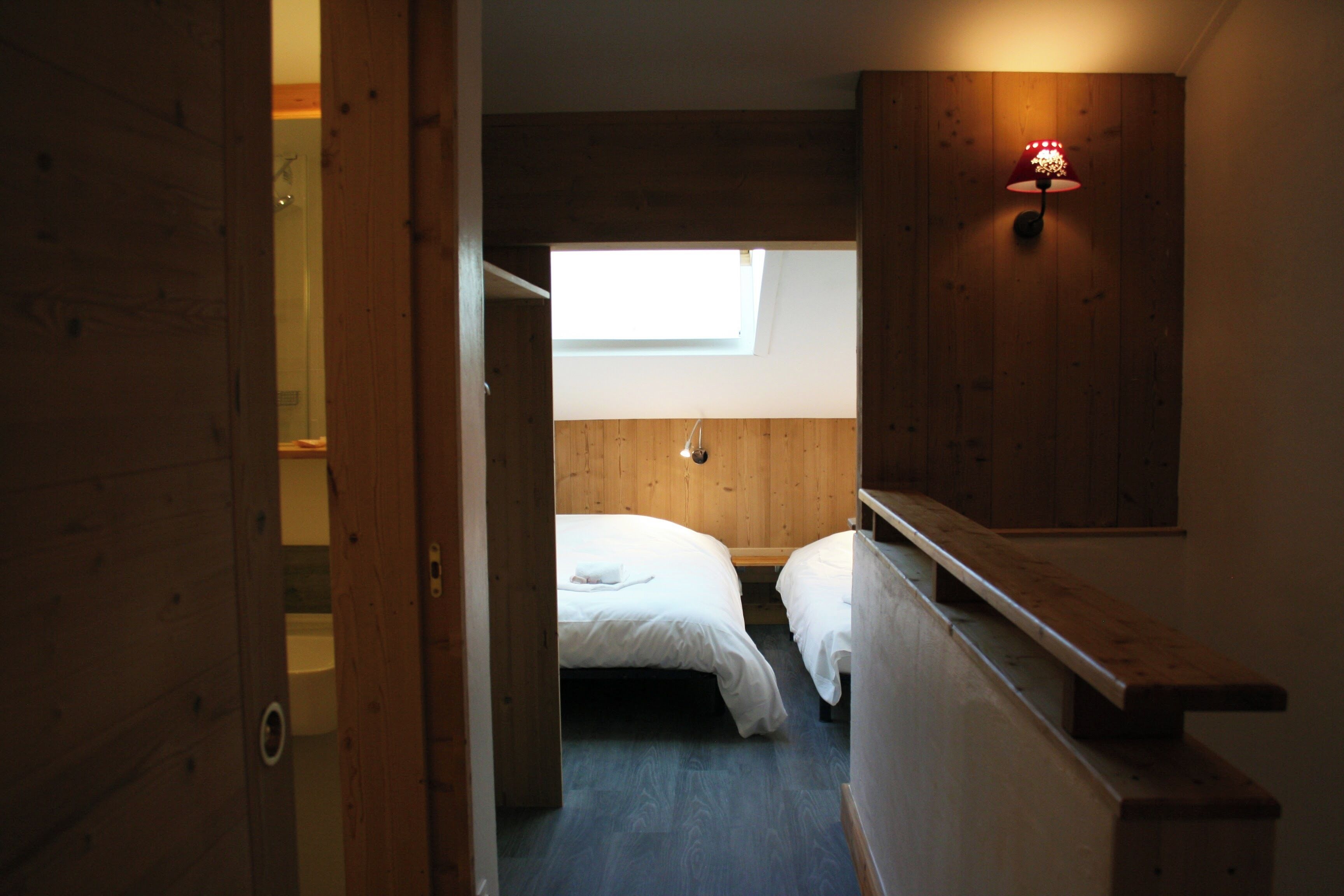 Een kleine, compacte kamer met een stapelbed en een enkel bed, houten wanden, een klein raam en een wandlamp.