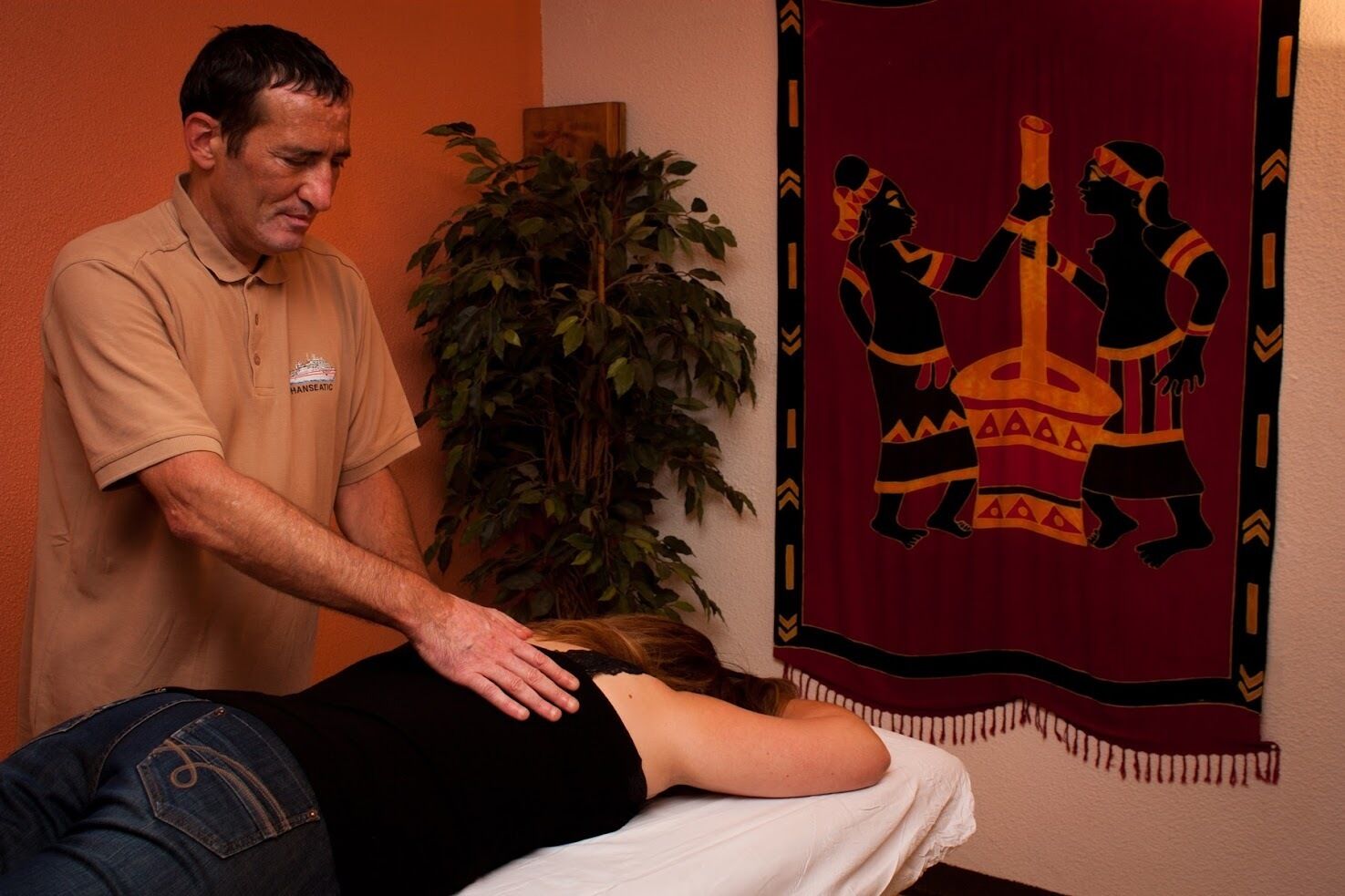 A massage therapy session in a room with a large plant and a tapestry depicting African figures.