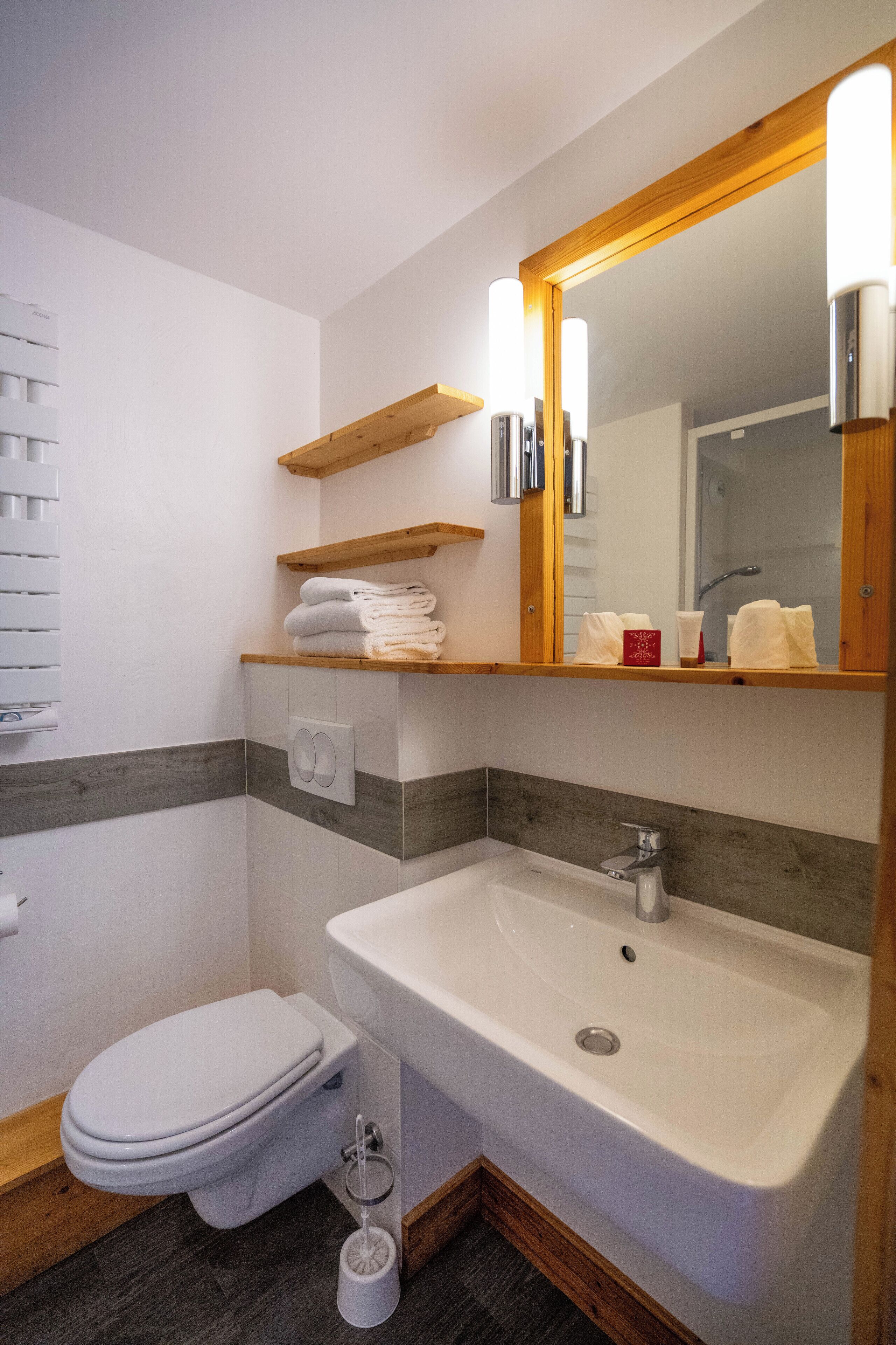 A modern bathroom with a white toilet, a sink, and a mirror. There are wooden shelves above the sink holding towels.