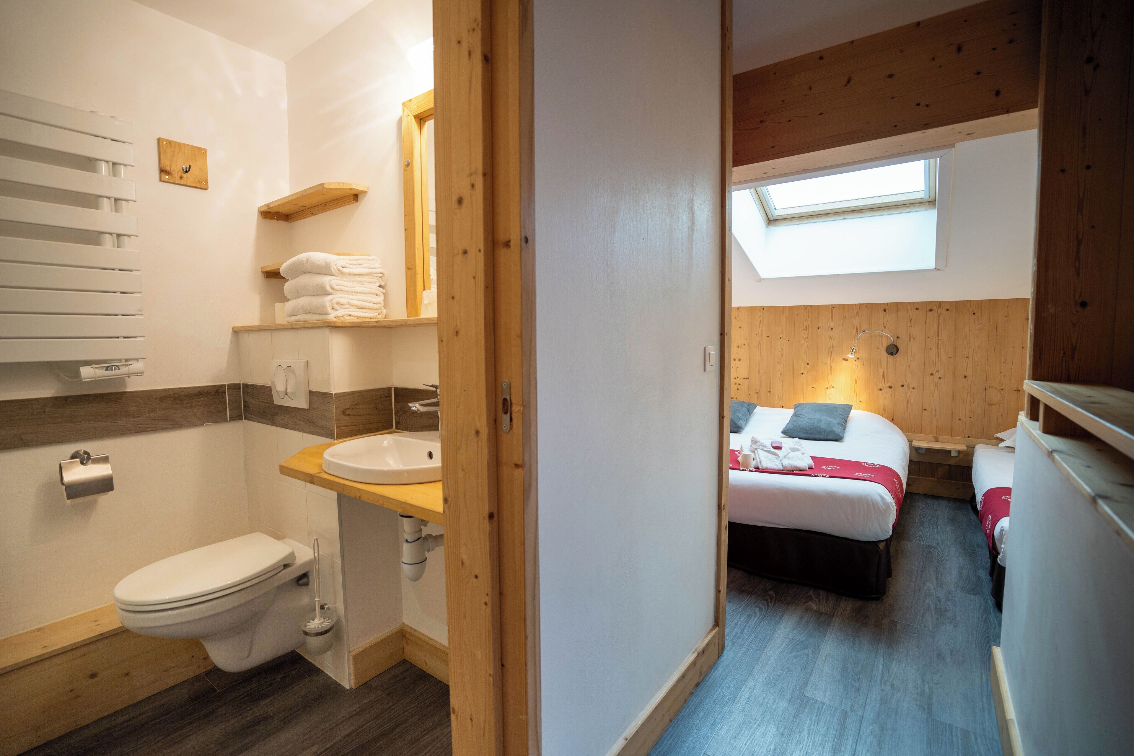 A compact bathroom with a toilet, sink, and wooden shelves. A skylight illuminates a bedroom with two beds.