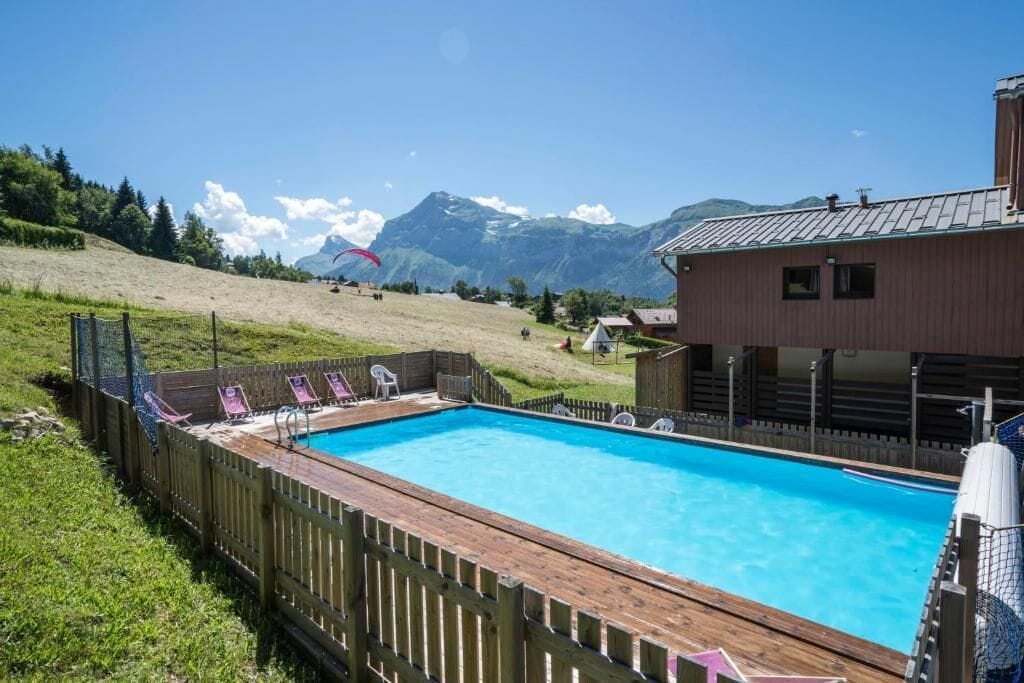 Une piscine dotée d’une terrasse en bois et de chaises longues, entourée d’une clôture en bois, avec des montagnes en arrière-plan.