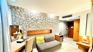 Una habitación de hotel moderna con una pared de detalles de piedra, un sofá con cojines, un escritorio de madera con una silla, una mesita de noche con una lámpara y un armario.