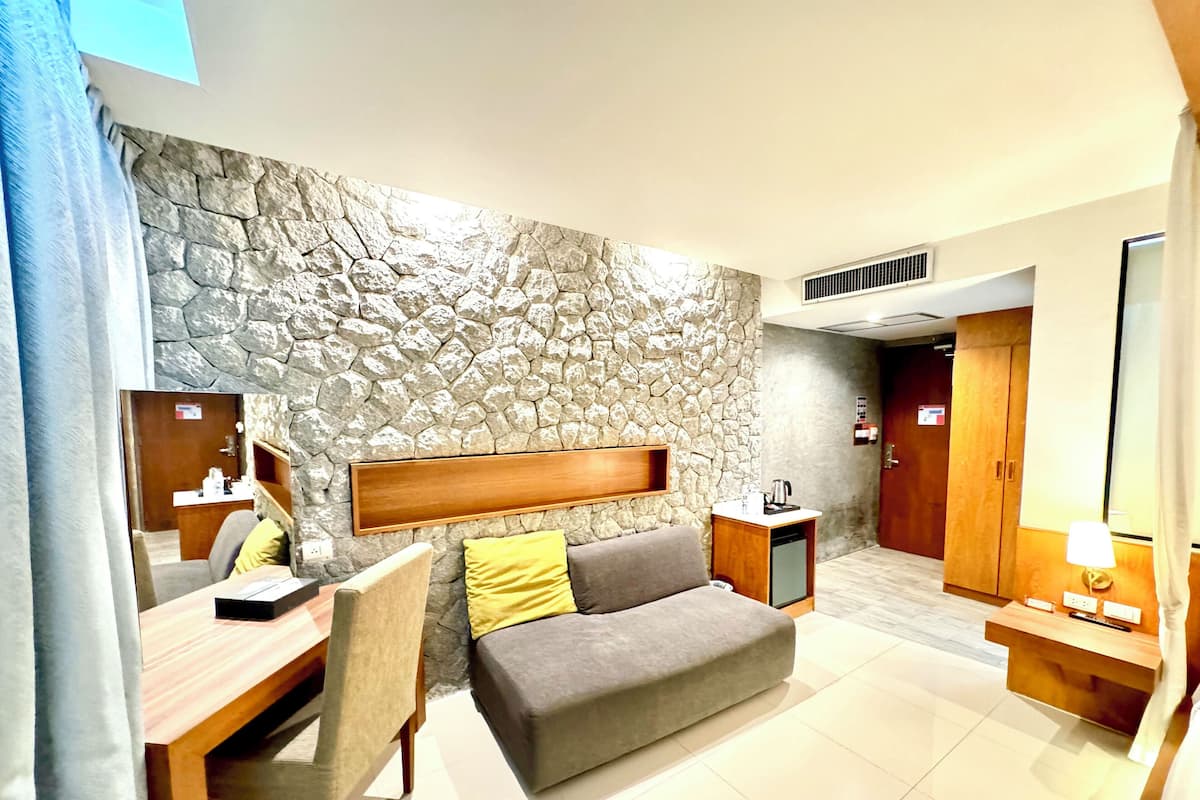 A modern hotel room with a stone accent wall, a sofa with cushions, a wooden desk with a chair, a bedside table with a lamp, and a wardrobe.