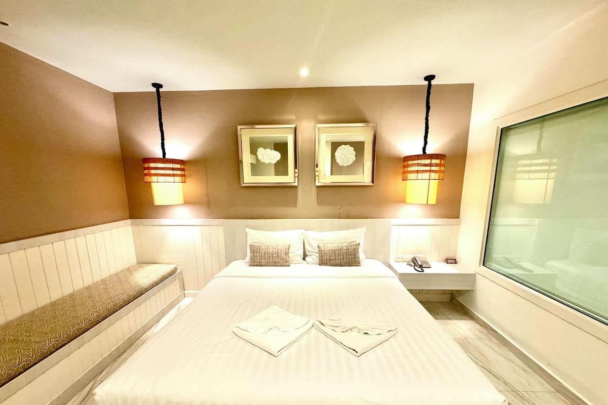 A hotel room with a large bed, two wall-mounted lamps, a bedside table with a lamp, a bench, and a window with a view.