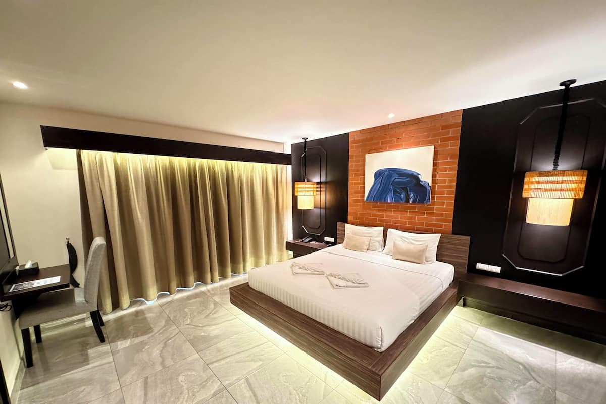 A modern hotel room with a large bed, a desk, and a chair. There is a window with curtains, a wall-mounted lamp, and a painting above the bed.