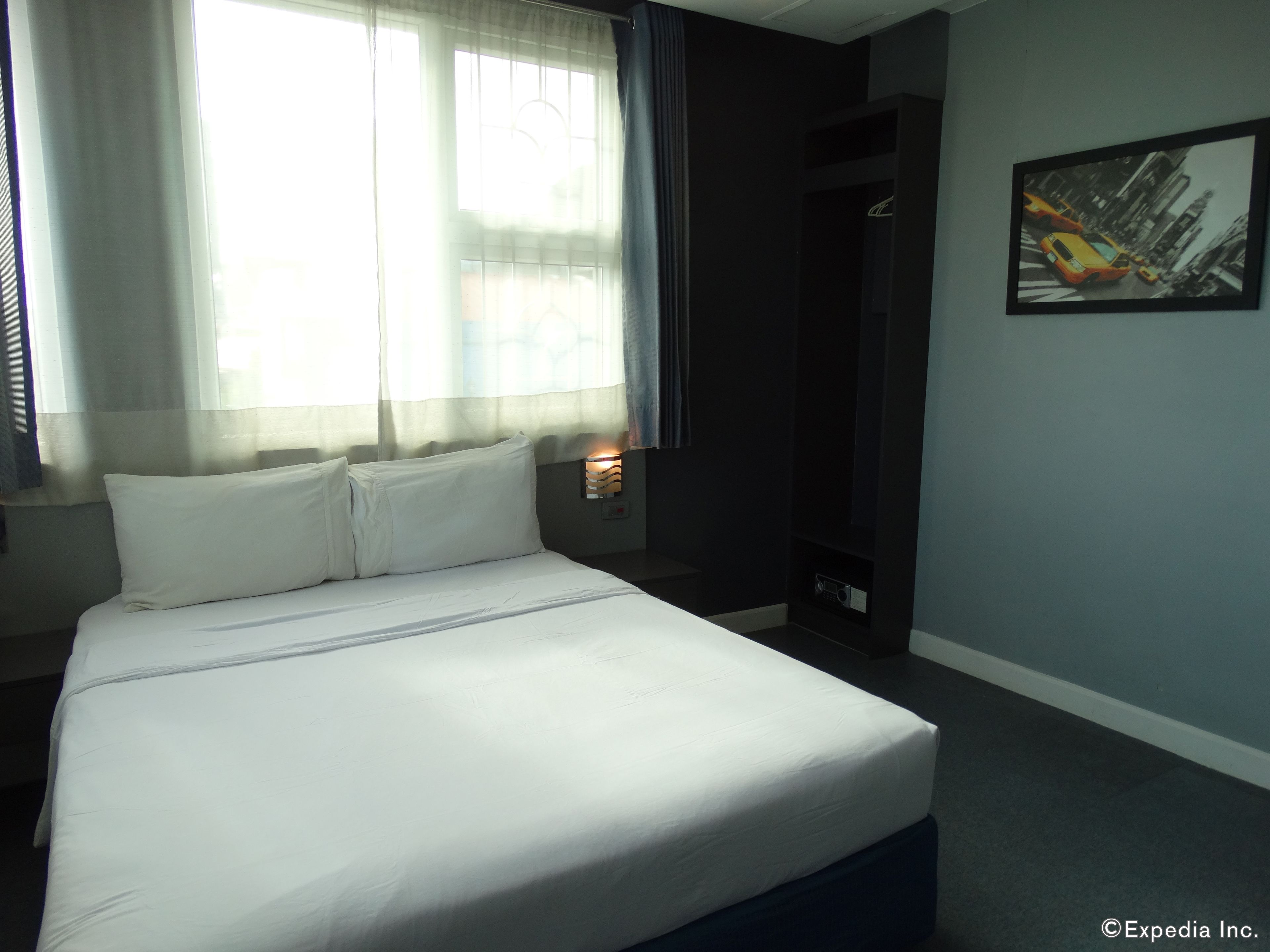 A hotel room with a bed, a window with sheer curtains, a painting on the wall, and a small table with a lamp.