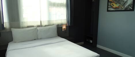 A hotel room with a bed, a window with sheer curtains, a painting on the wall, and a small table with a lamp.