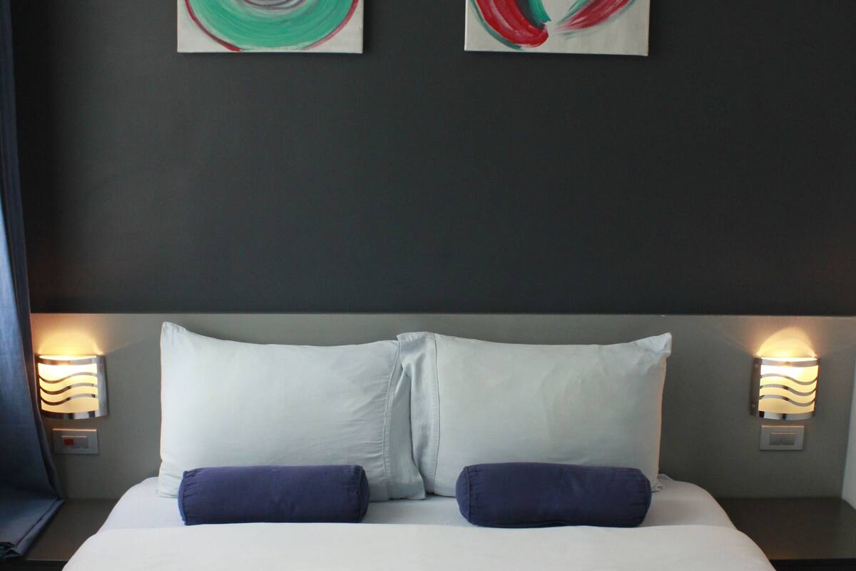 A neatly made bed with white and blue pillows, two wall-mounted lamps, and two abstract paintings on the dark wall above.