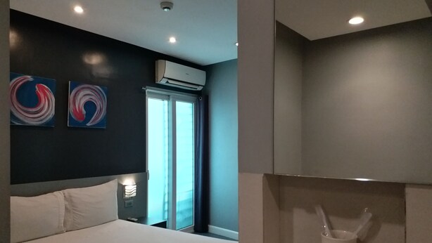 A modern hotel room with a large bed, two abstract wall art pieces, an air conditioning unit, a window with curtains, and a small white cabinet with a spoon and cup. - Leez Inn Malate (Manila)