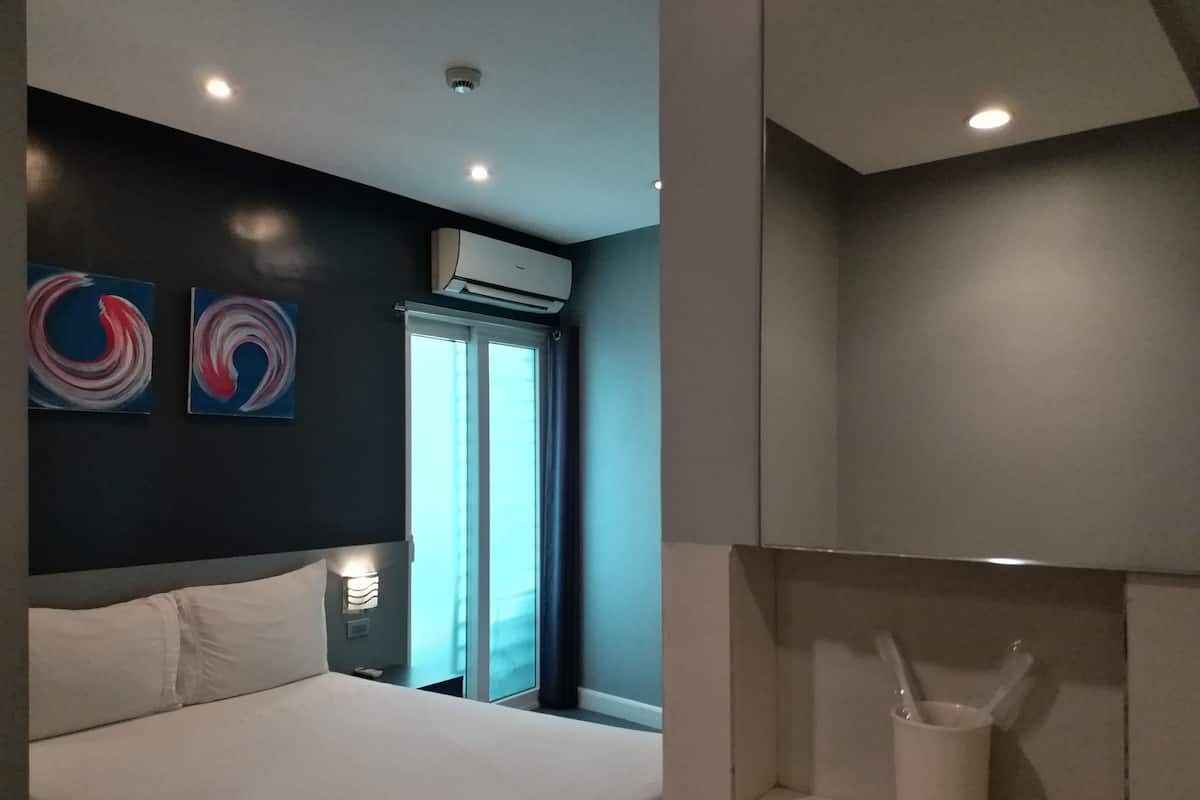A modern hotel room with a large bed, two abstract wall art pieces, an air conditioning unit, a window with curtains, and a small white cabinet with a spoon and cup.
