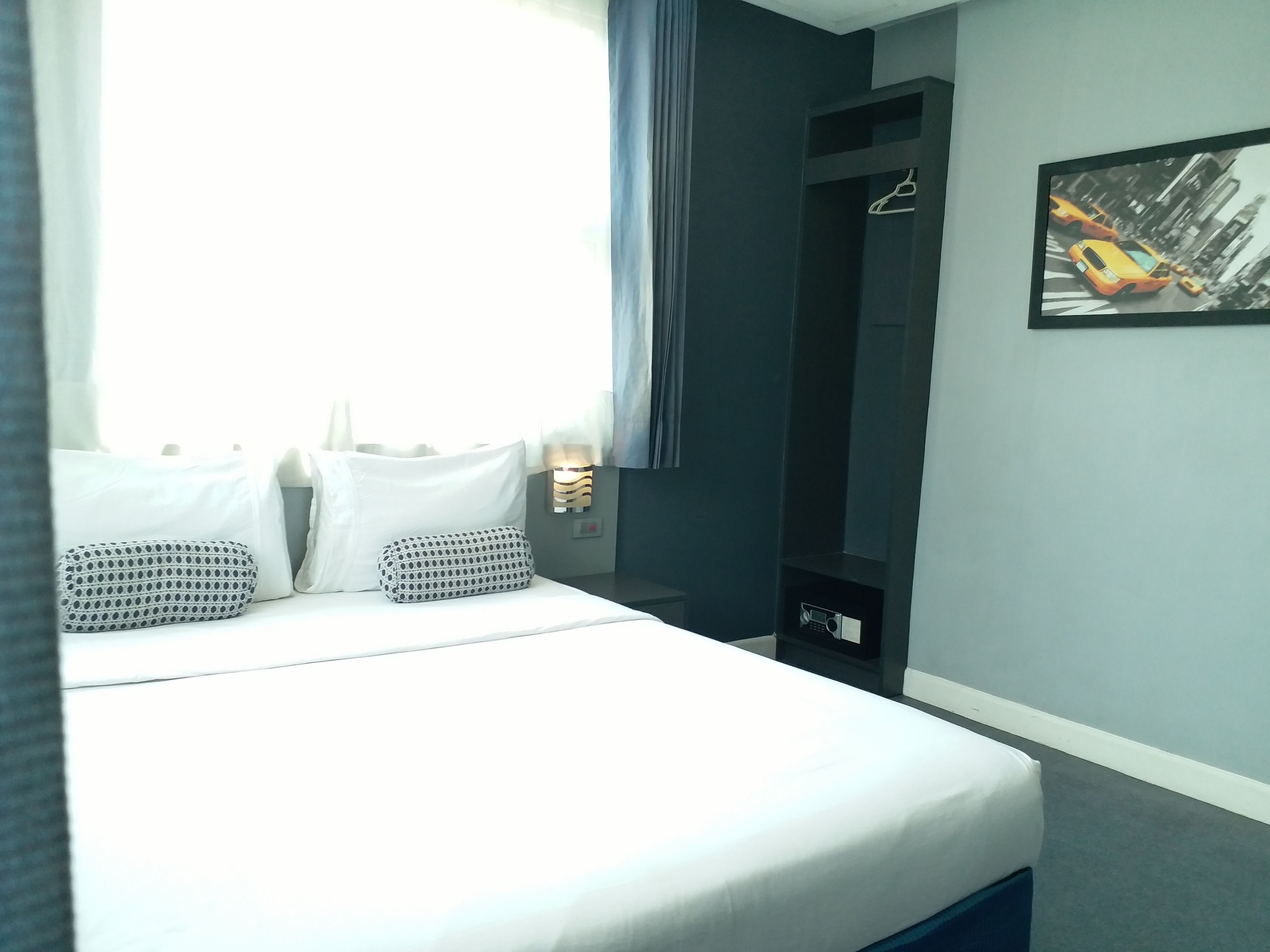 Een net opgemaakt bed met witte lakens en gekleurde kussens, een nachtkastje met een lamp en een kapstok aan de muur.