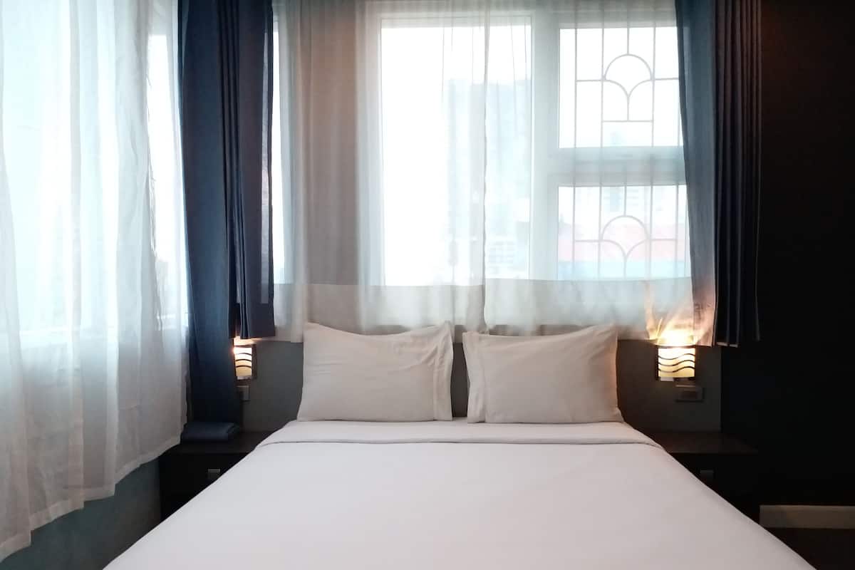 A neatly made bed with white linens in a room with a large window, sheer curtains, and wall-mounted lamps.