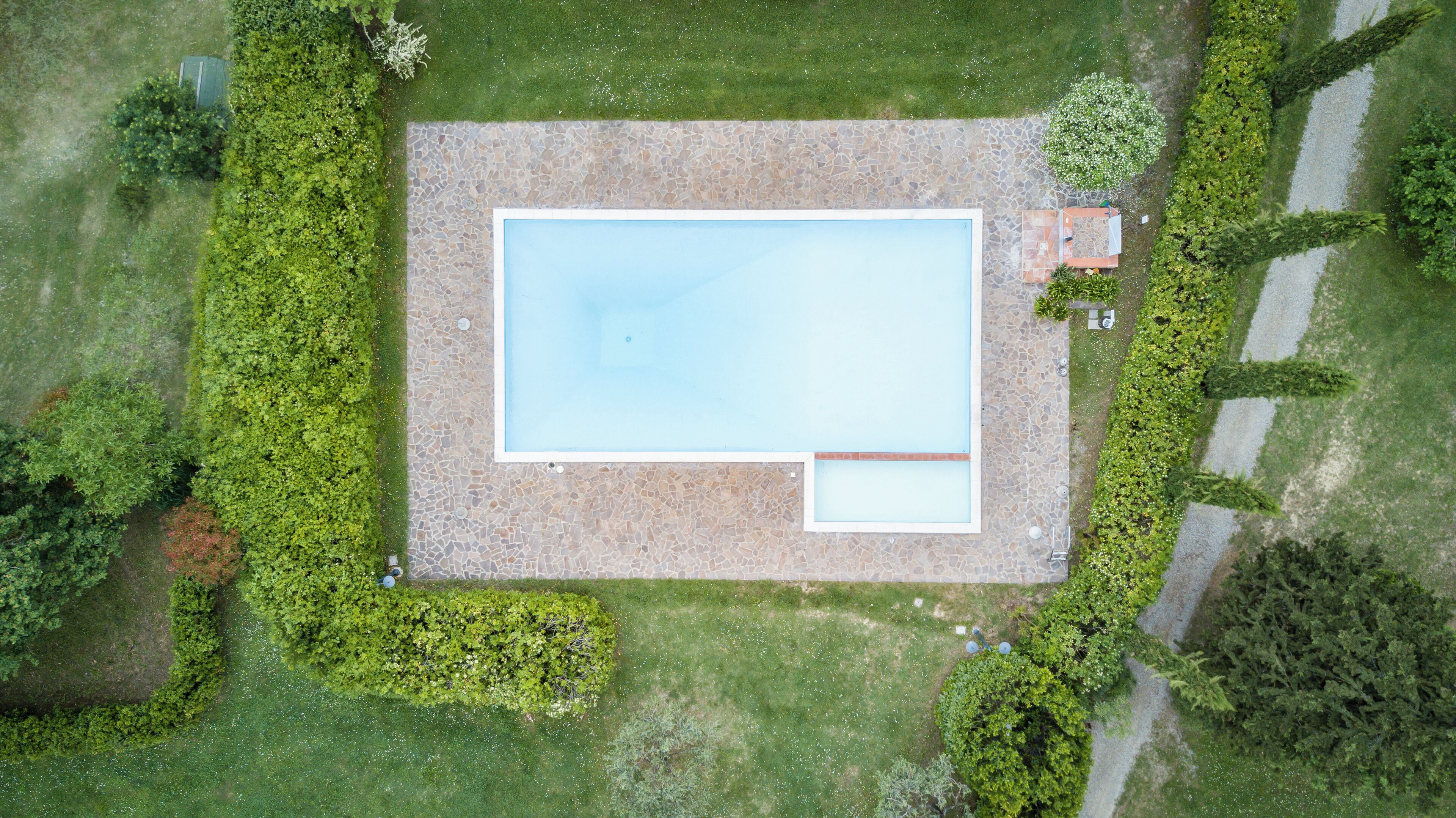 Ovenudblik over et rektangulært swimmingpool med flisebelagte kanter, omgivet af velplejede græsplæner og hække.