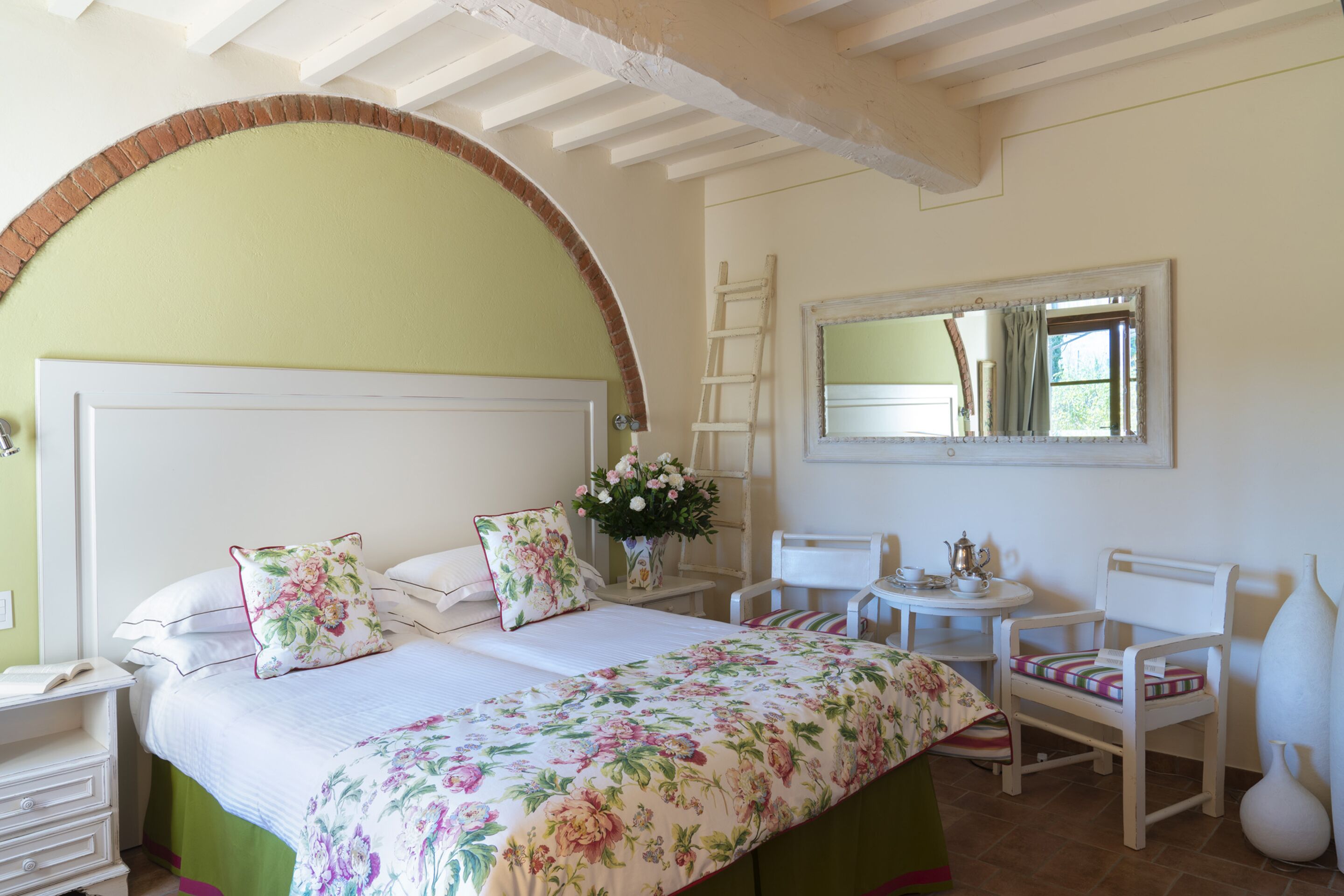 A bedroom with a large bed, a bedside table, a chair, a small table with a teapot, and a mirror on the wall.