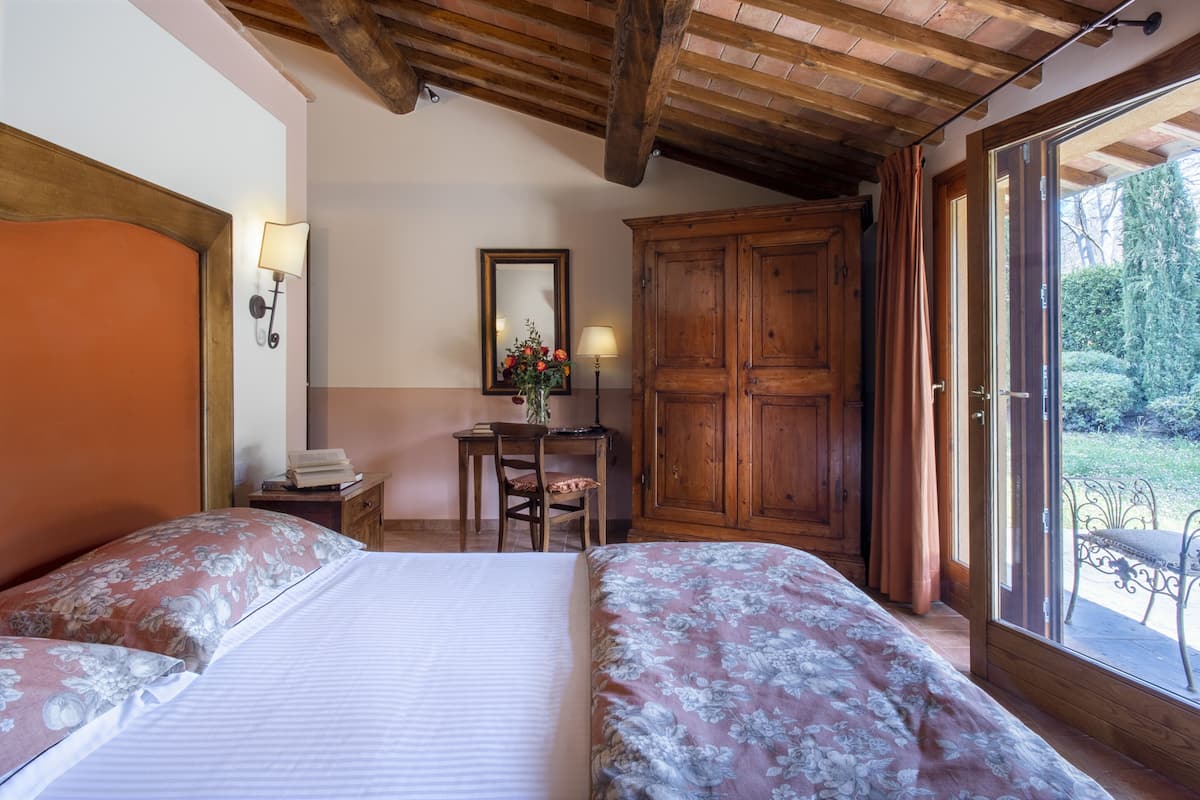 Quarto com duas camas, um guarda-roupa de madeira, uma mesa de trabalho com cadeira, espelho e janela com cortinas.