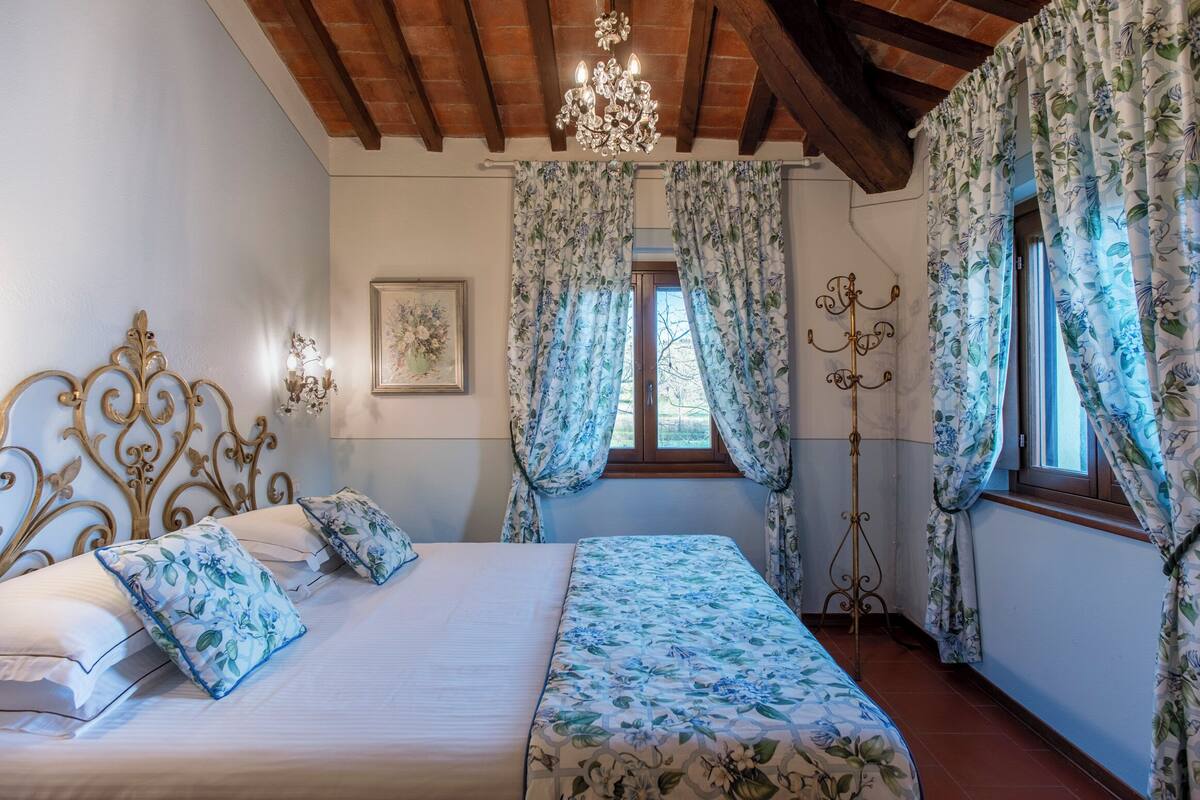 Quarto com cama, cortinas florais, um candelabro e um abajur.