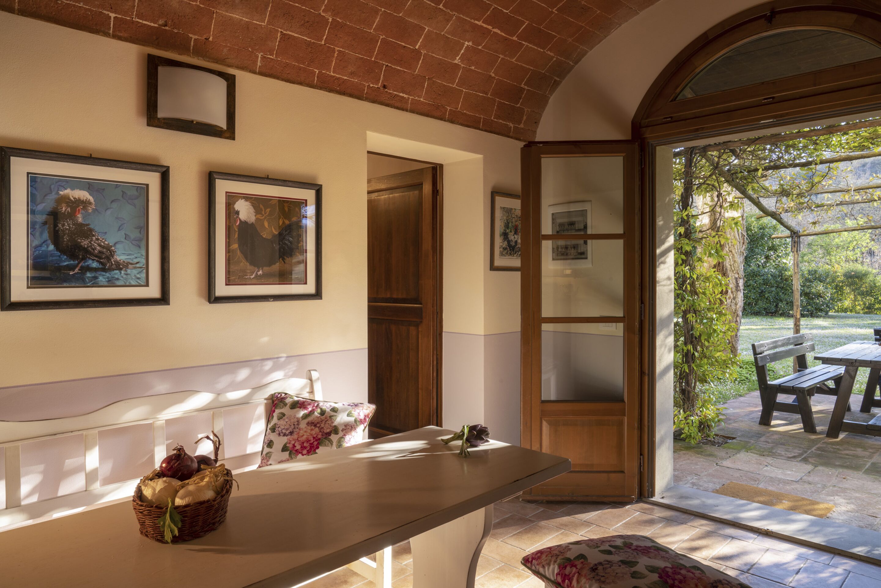 A reception area with a counter, framed artwork, a basket of fruits, and a view of an outdoor patio with a table and chairs.