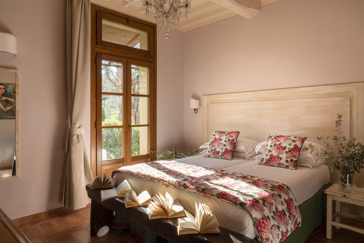 Quarto com uma cama grande, um banco de madeira com livros, uma janela com cortinas e um lustre.