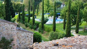 A stone building with a garden featuring tall trees, a swimming pool, and lounge chairs with blue umbrellas. - Borgo san Benedetto (Montaione)