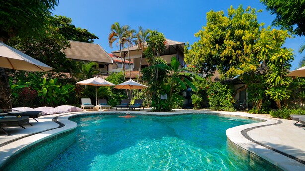 A swimming pool with lounge chairs and umbrellas, surrounded by lush greenery and buildings. - Pearl Boutique Hotel - Adult Only (Legian)