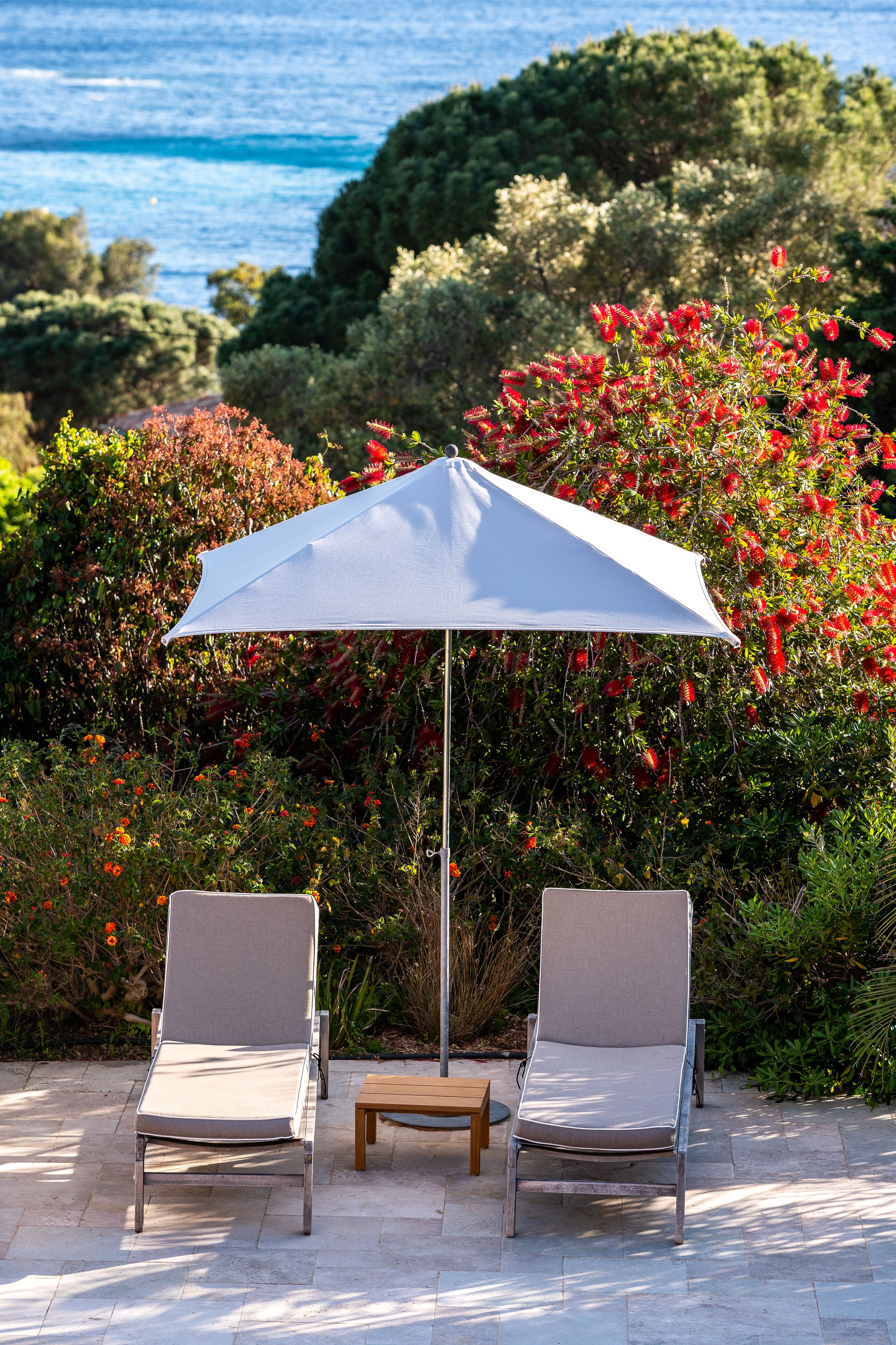 A patio with two lounge chairs and a table under a white umbrella, surrounded by lush greenery and a view of the sea.