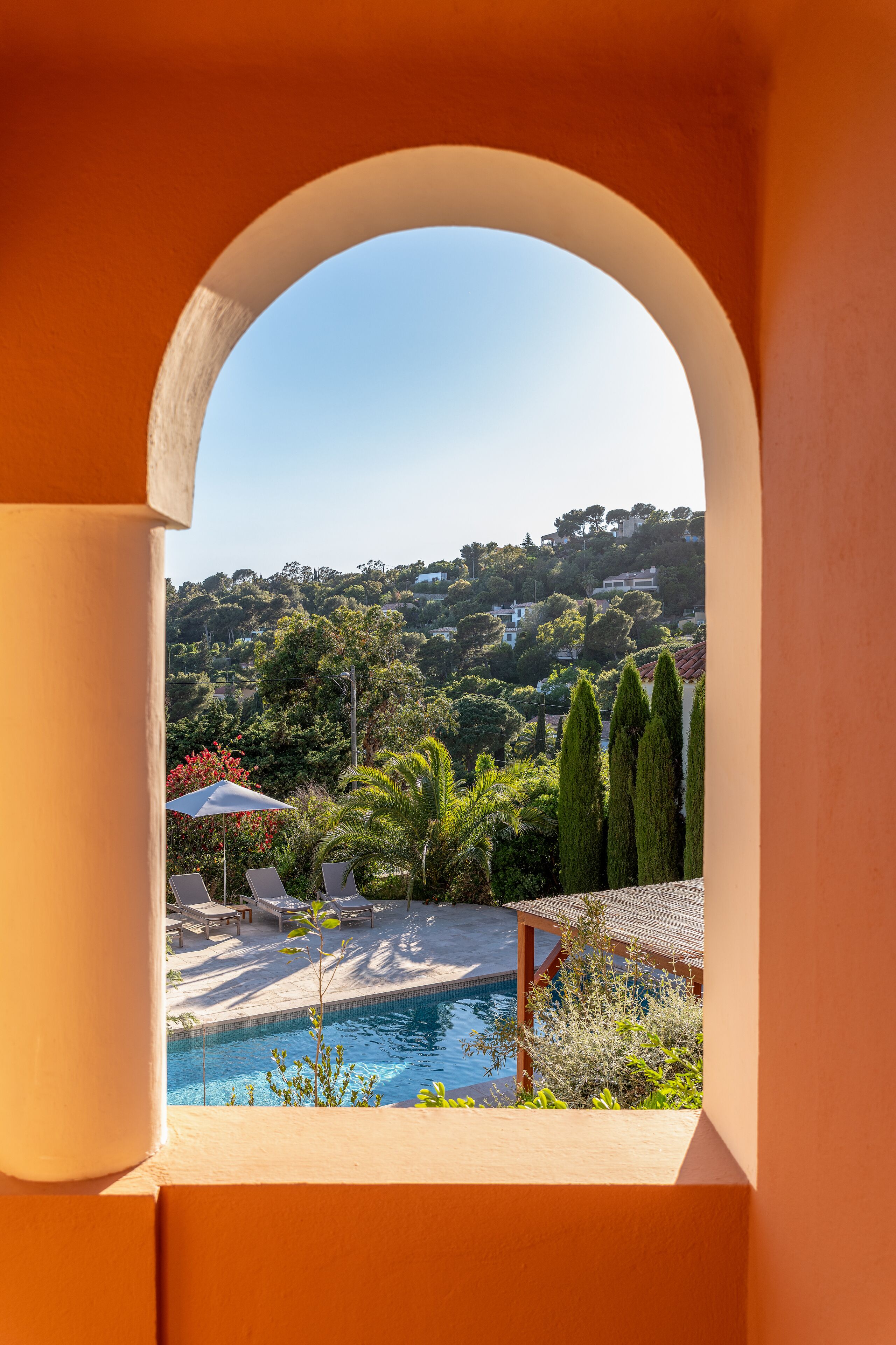 Blick durch einen Bogen auf einen Poolbereich mit Liegestühlen, einem Tisch und einer Aussicht auf einen Hügel mit Bäumen und Häusern.
