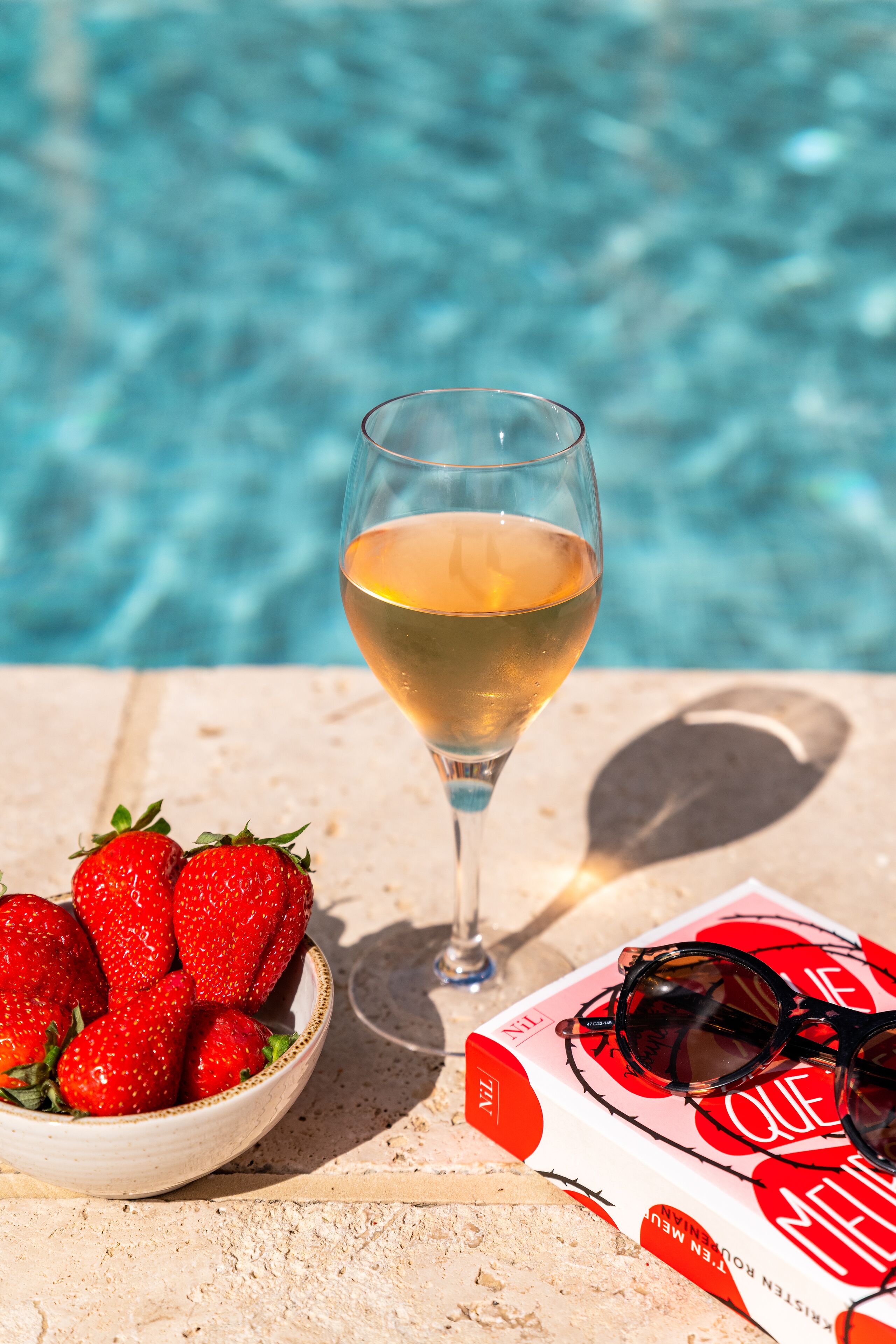 Una copa de vino blanco, un cuenco de fresas y una revista con gafas de sol sobre una mesa junto a una piscina.