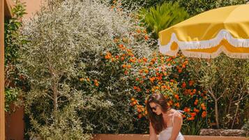 A woman sitting on a sun lounger in a courtyard with potted plants and a yellow umbrella.