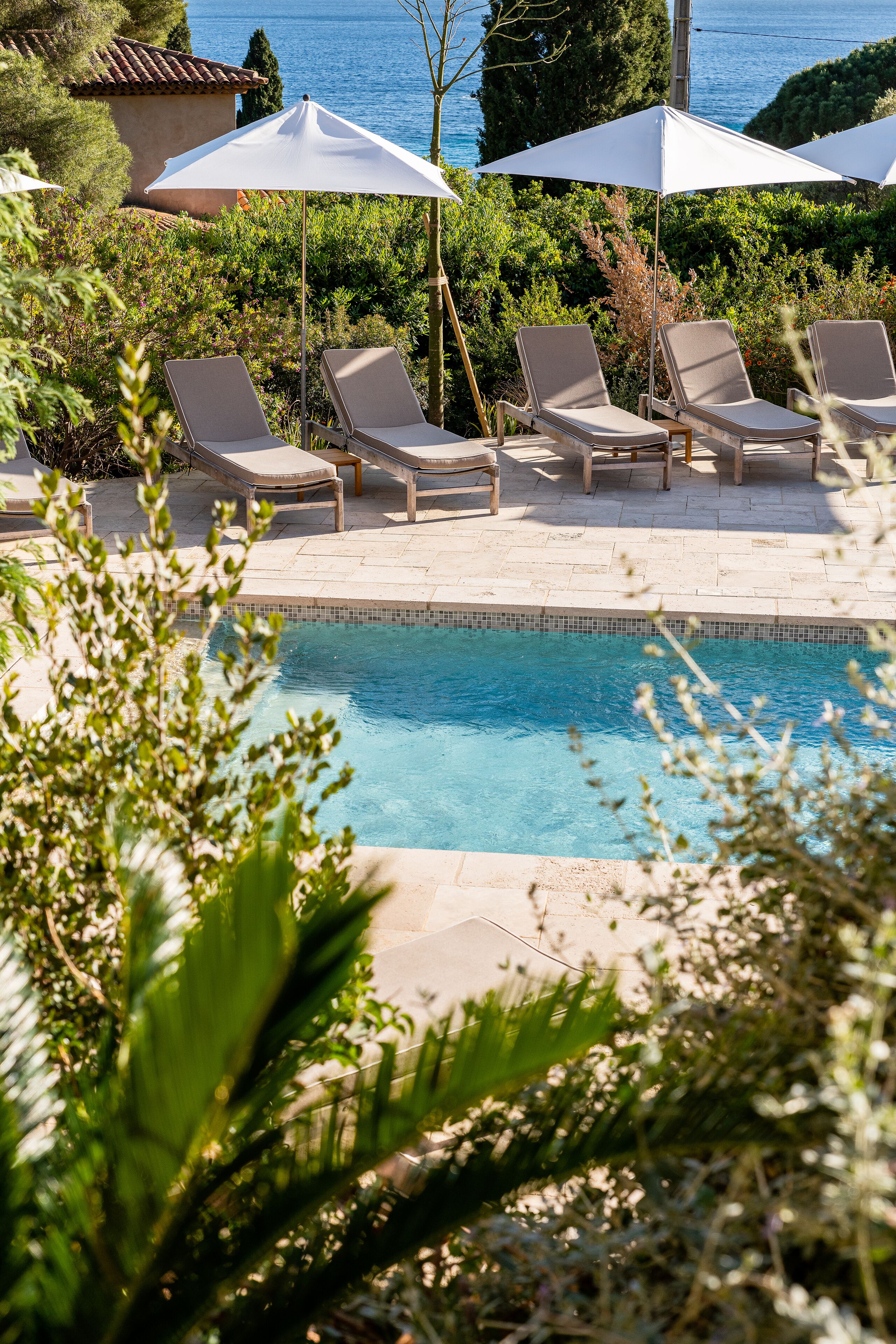 A pool area with lounge chairs and umbrellas, surrounded by greenery and overlooking a body of water.
