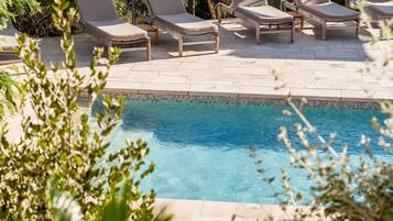 A pool area with lounge chairs and umbrellas, surrounded by greenery and overlooking a body of water.