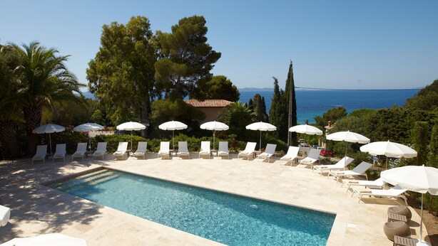 Un espace piscine agrémenté de chaises longues et de parasols, entouré d’arbres et offrant une vue sur la mer.