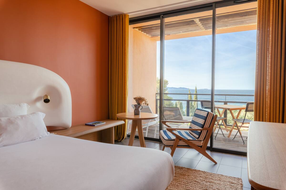 A hotel room with a bed, a small table, a chair, and a balcony with a view of the sea.