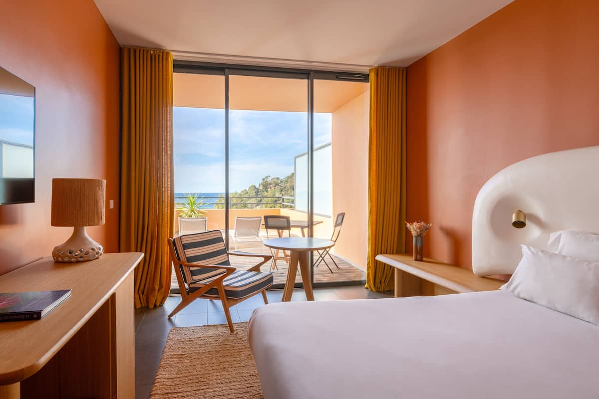 A hotel room with a bed, a desk, a chair, a small table, and a view of the outdoors.