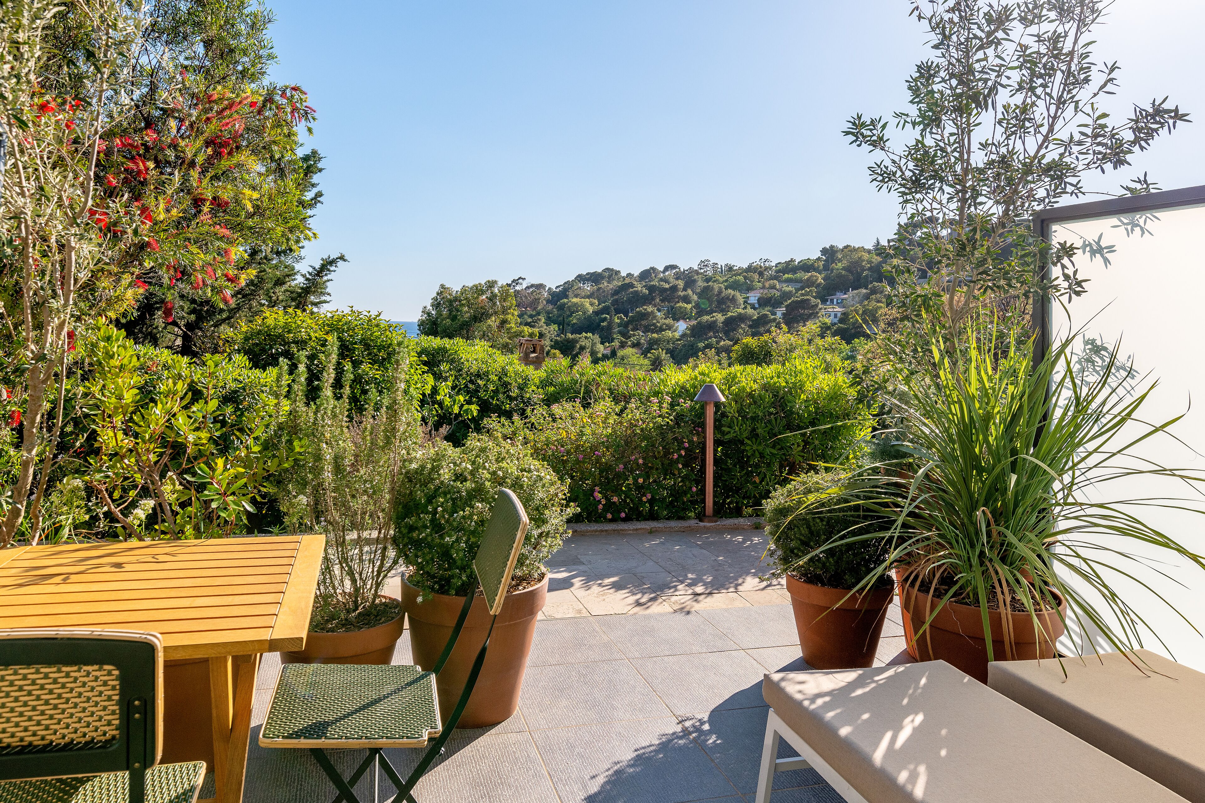 Un patio exterior con una mesa y sillas de madera, rodeado de plantas en macetas y vistas a un jardín exuberante y una colina al fondo.