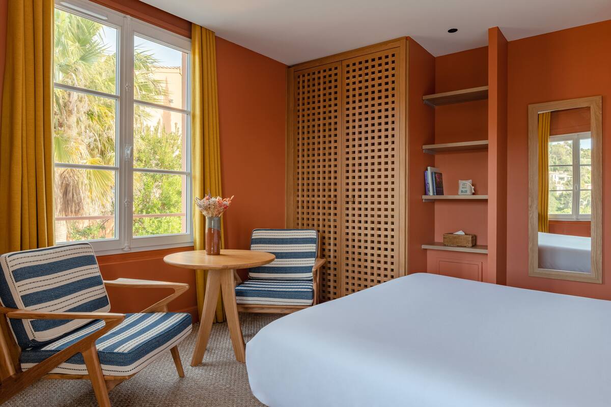 A hotel room with a large bed, two striped chairs, a small round table with a vase, and a wooden louvered partition.