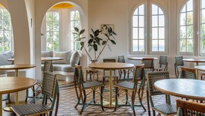 Un espace repas avec des tables rondes et des chaises en osier, un canapé et de grandes fenêtres donnant sur la verdure.