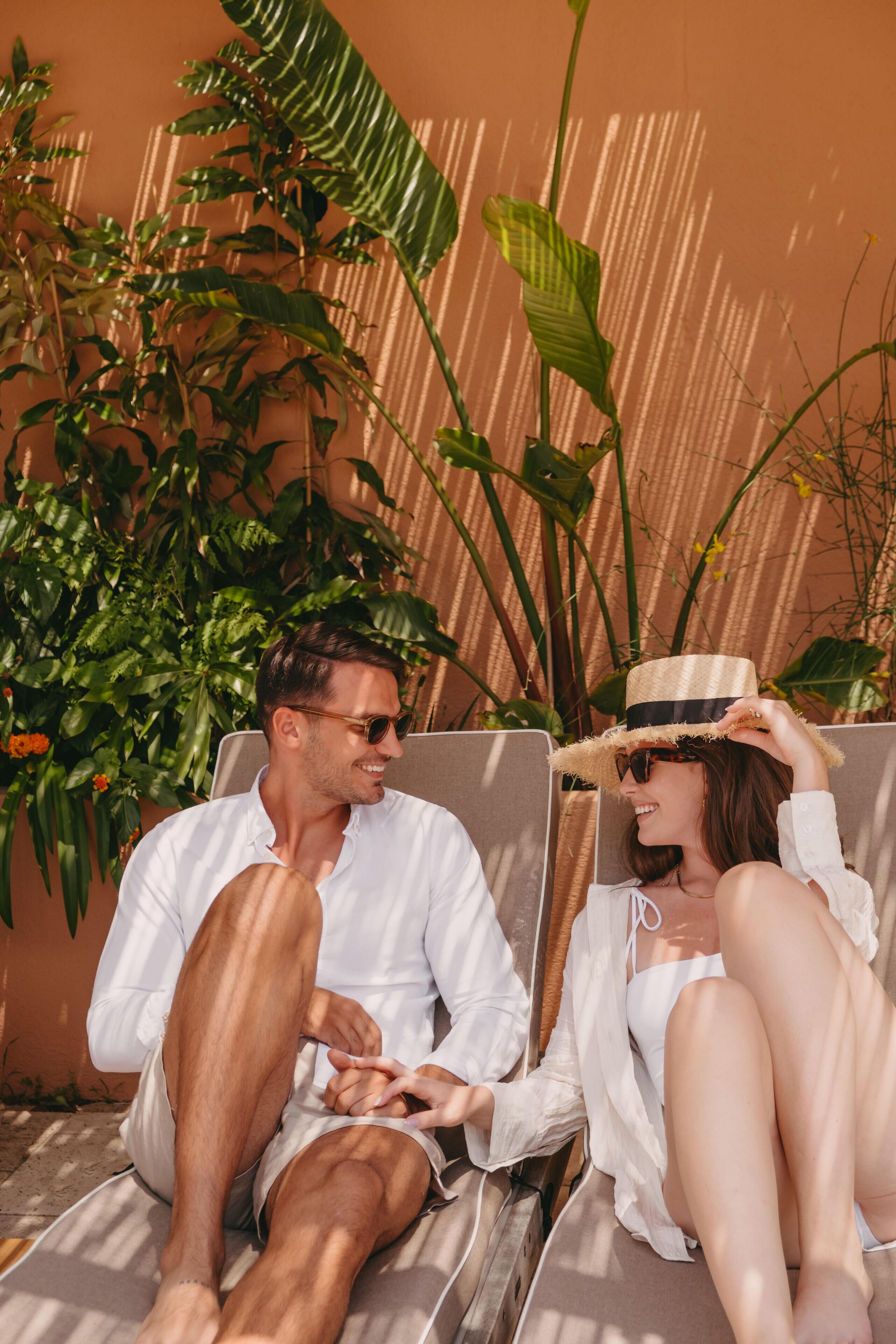 Two people sitting on sun loungers with a plant and a wall in the background.