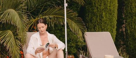 A woman sitting on a sun lounger under a yellow umbrella, reading a book.