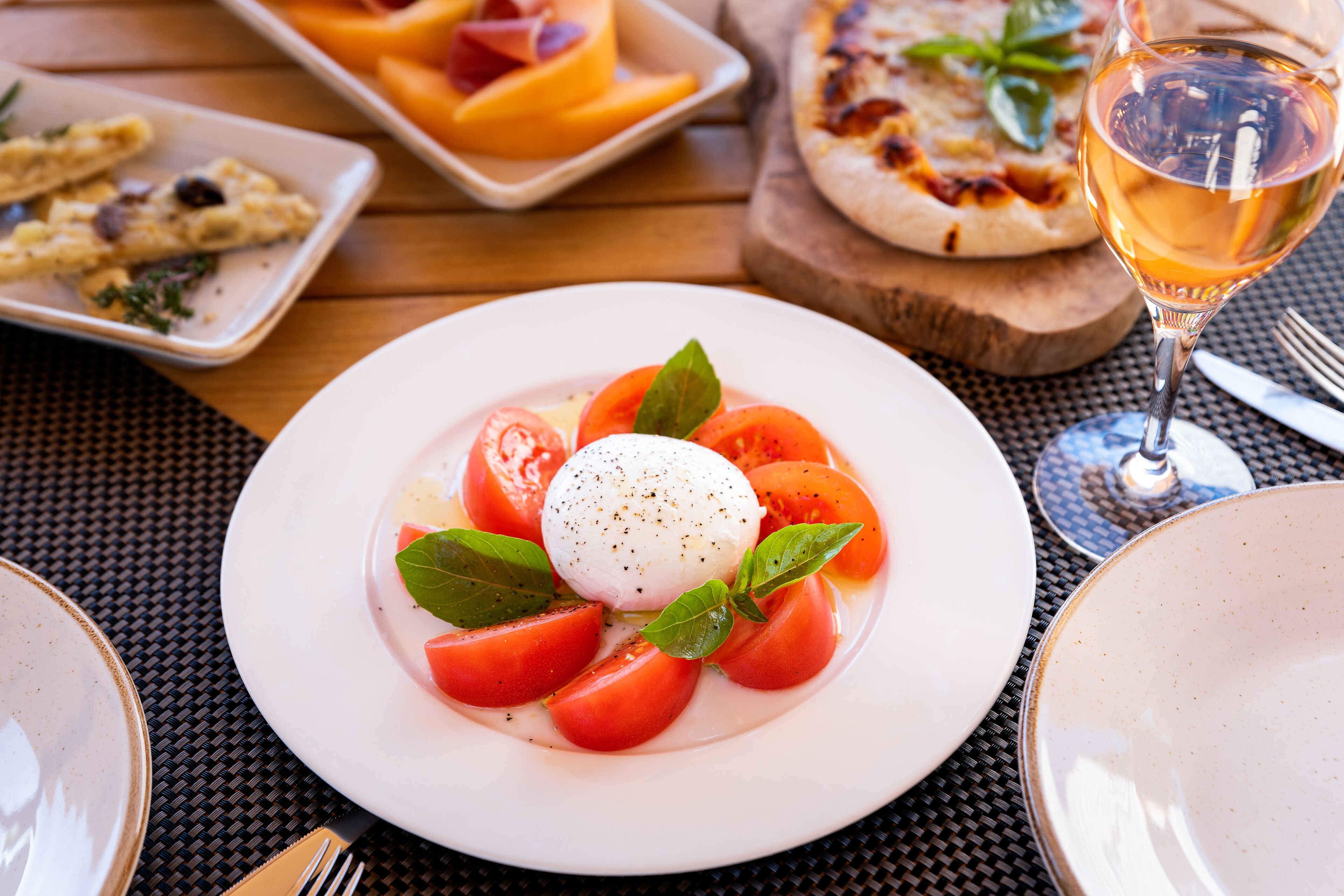 Une assiette de salade caprese, composée de tomates, de mozzarella et de basilic, est posée sur une table. On aperçoit en arrière-plan une pizza et un verre de vin.